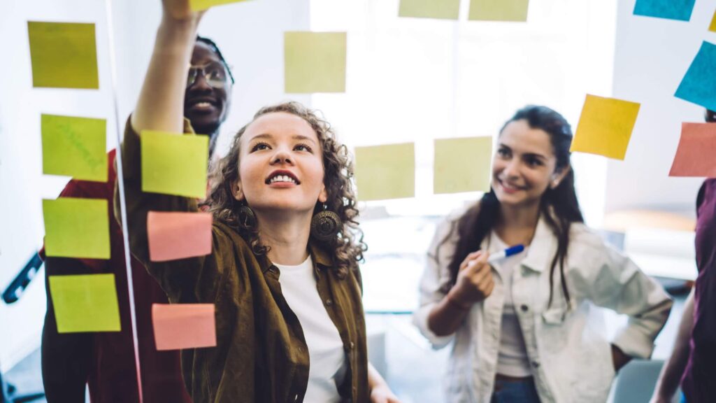 A diverse team of three young entrepreneurs standing in front of a glass wall covered in colorful sticky notes, brainstorming and collaborating on a strategy roadmap in a bright office space.