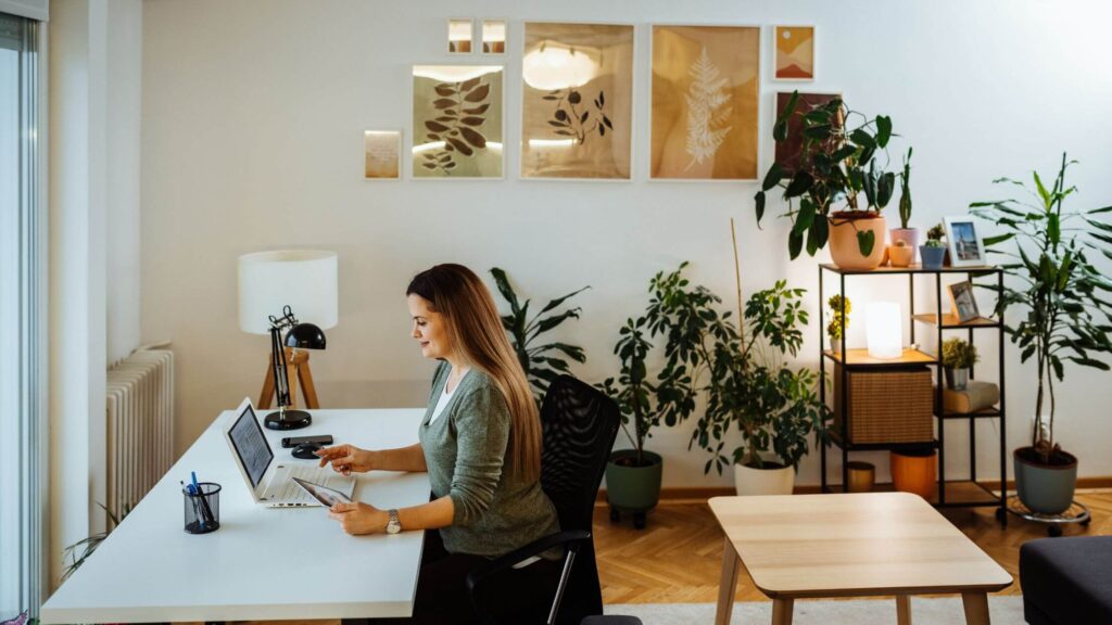 A woman sitting at a white desk in a bright, modern office with plants and warm artwork on the walls. She is working on a laptop, with an open notebook, pens, and a mug nearby, creating a professional and focused environment.