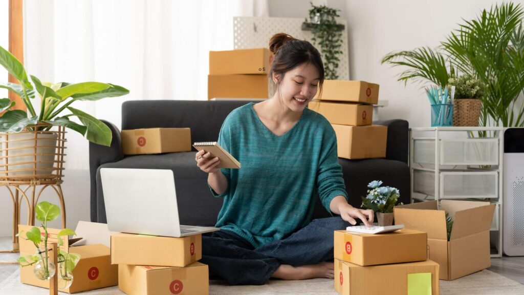A smiling woman sitting cross-legged on the floor of a bright living room, surrounded by cardboard boxes ready for shipping. She is holding a tablet, looking at an open laptop on a low table, and managing her e-commerce business operations.